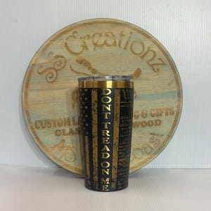 May include: A black and gold tumbler with the text "DON'T TREAD ON ME" in gold lettering. The tumbler features a design of the American flag and a gold rim. A wooden sign is in the background.
