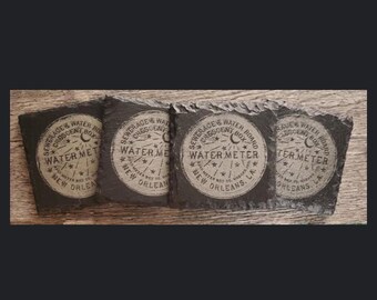 May include: Four square, dark gray slate coasters. Each coaster features a circular design with the text "SEWERAGE & WATER BOARD CRESCENT BOX WATER METER NEW ORLEANS, LA" in a vintage style. The design includes a crescent moon and stars.