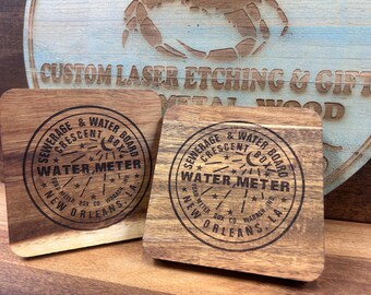 May include: Two square wooden coasters with rounded corners. Each coaster has a circular black graphic with the words "SEWERAGE & WATER BOARD CRESCENT BOX WATER, METER NEW ORLEANS, LA" etched into the wood. A wooden sign is in the background.