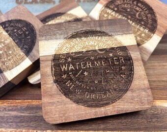 May include: Wooden square coasters with a two-tone wood grain design. Each coaster features a circular engraved design with the words "Sewerage & Water Board Crescent Box Water Meter New Orleans".