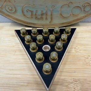 May include: A triangular wooden game board with a black top surface featuring fourteen brass-colored bullet casings arranged in a pattern. The board has a small circular hole in the center and is positioned in front of a curved wooden sign with the text Gulf Coast.