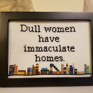 Op de afbeelding: Een ingelijst kruissteek met de tekst "Dull women have immaculate homes." Het kruissteek is wit met zwarte tekst en een rij kleurrijke boeken aan de onderkant.