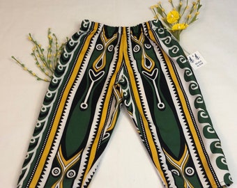 May include: Green, yellow, black, and white patterned capri pants. The pants have an elastic waistband and a unique tribal design. The fabric is lightweight and suitable for warm weather. The pants are displayed on a white surface.