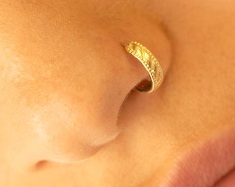 May include: A gold nose ring with a decorative design. The ring is worn in the left nostril.