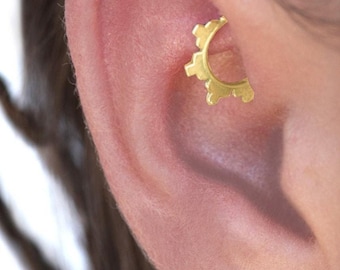 May include: A gold-colored ear piercing in the shape of a gear. The jewelry is placed in the upper ear cartilage. The image shows a close-up of the ear and the piercing.