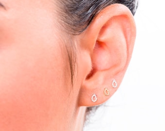 May include: Close-up of an ear with three small, teardrop-shaped earrings. Two are silver with clear stones, and one is gold. The earrings are delicate and minimalist, suitable for everyday wear.