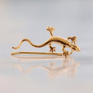 May include: A gold-colored lizard-shaped hair clip. The lizard is depicted in a stylized form, with a long tail and detailed legs. The clip is designed to be worn in the hair and is made of a shiny, metallic material.