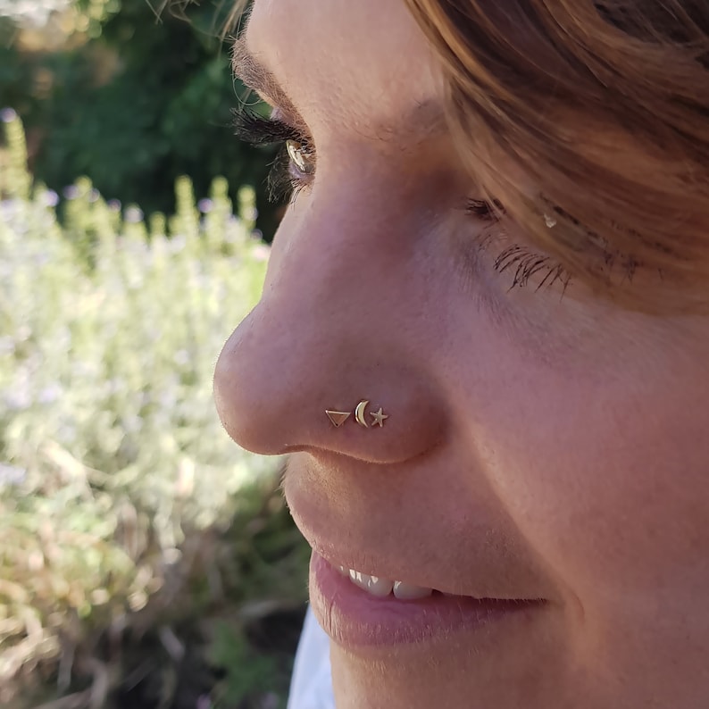May include: Close-up of a person's nose with a small gold triangle, crescent moon, and star nose ring.