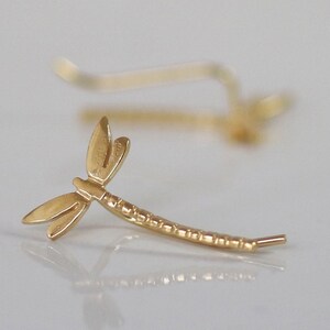 May include: A gold-colored dragonfly ear climber earring. The earring features a detailed dragonfly design with delicate wings and a textured body. The earring is designed to curve along the ear, creating a unique and elegant look.