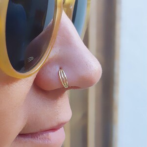 May include: A close-up of a person's nose with a gold nose ring. The ring is made of three thin bands of metal.
