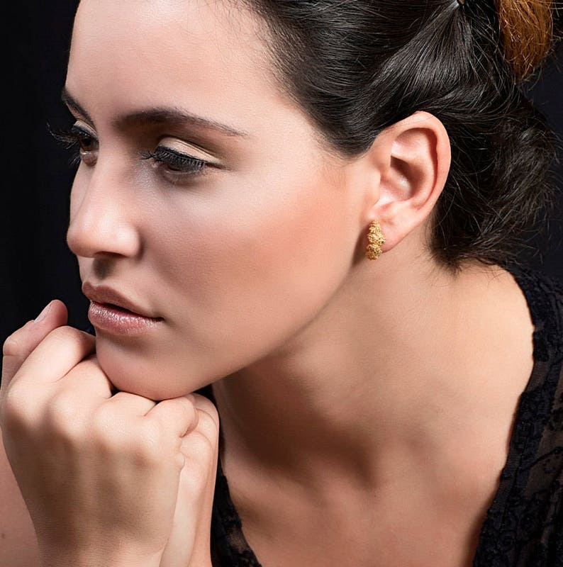 May include: A woman with dark hair and a thoughtful expression is wearing gold hoop earrings. The earrings have a textured, floral design.