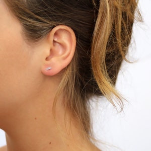 Puede incluir: Primer plano de la oreja de una persona con un pequeño pendiente de plata en forma de pluma. El pendiente tiene un diseño sencillo con una curva delicada.