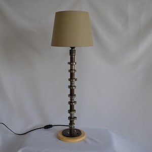May include: A table lamp with a beige fabric lampshade and a base made from a metal engine part. The base is mounted on a wooden platform.