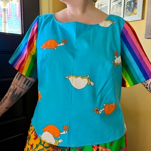 May include: A turquoise blue shirt with rainbow striped sleeves and a pattern of orange and white turtles carrying flowers. The shirt has a scoop neck and is made of a soft, cotton fabric.