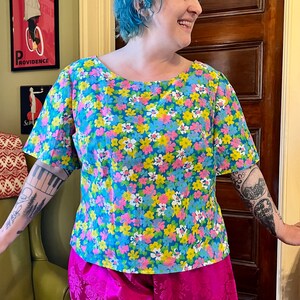 May include: A floral print top with a scoop neck and short sleeves, paired with bright pink pants. The top features a pattern of blue, yellow, and pink flowers on a light blue background. The pants have a floral pattern.