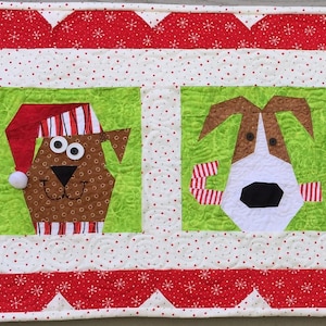 Pode incluir: Uma tapeçaria de parede acolchoada com tema natalino com quatro cães usando chapéus de Papai Noel. Os cães são marrons, brancos, preto e branco e pretos. O fundo é branco com detalhes vermelhos e verdes.