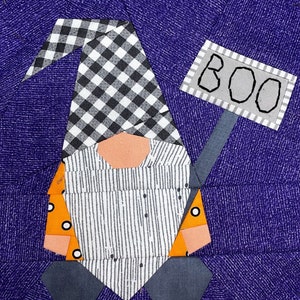 Peut inclure: Un gnome en patchwork tenant une pancarte sur laquelle est inscrit "BOO" en lettres noires sur fond gris. Le gnome porte un chapeau à carreaux noirs et blancs, une chemise rayée et un pantalon orange. L'arrière-plan est un tissu violet à motif texturé.
