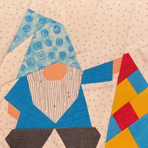 May include: A fabric patchwork block featuring a whimsical gnome design. The gnome has a blue hat with a swirl pattern, an orange face, and a blue jacket. The background is a light fabric with small gray polka dots. The gnome's beard is white with vertical gray stripes.