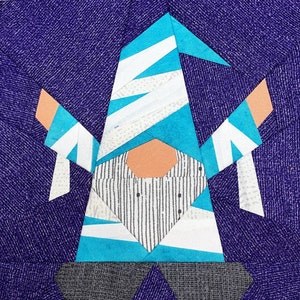 Op de afbeelding: Een blauwe en witte patchwork-gnome met een gestreept shirt en een witte muts. De gnome staat op een paarse achtergrond.
