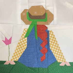 May include: A patchwork quilt block depicting a girl with a brown hat, green ribbon, red braid, and a blue dress. The girl is standing on a green patch of grass with a pink flower in the background.