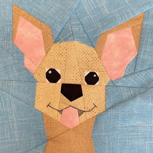 May include: A fabric art piece featuring a geometric dog design. The dog's face is constructed from various fabric pieces in tan, pink, and black, with a light blue background. The dog has black eyes, a pink tongue, and a smiling mouth.