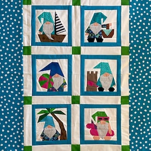May include: A white quilt with a blue and green border. The quilt is divided into six squares, each featuring a different gnome with a summer theme. The gnomes are wearing different outfits and accessories, such as a boat, a beach umbrella, a sandcastle, a palm tree, and a float.