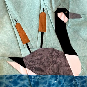 May include: A patchwork quilt block depicting a stylised image of a goose swimming in blue water. The goose is made from black, white and grey fabric. The water is made from blue fabric with a swirling pattern. The goose has two brown reeds sticking up from its back.