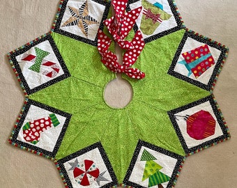 May include: A green quilted Christmas tree skirt with a white, black, and red patchwork design. The skirt features various holiday-themed motifs, including candy canes, ornaments, and a star.