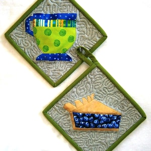 Op de afbeelding: Twee gewatteerde pannenlappen. Eén heeft een groene beker met blauwe en gele accenten. De andere toont een stuk bosbessentaart. Beide zijn vierkant met een groene rand en een lus om op te hangen.