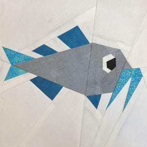May include: A gray and blue patchwork fabric fish with a black eye. The fish has a white background and is made of different fabric pieces.