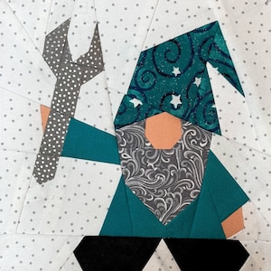May include: A patchwork gnome design featuring a teal tunic, a patterned hat with stars, and a gray beard holding a wrench. The gnome's face is peach-coloured, set against a white background with gray polka dots.