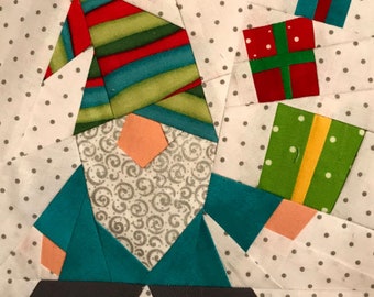 Puede incluir: Un bloque de edredón de tela que muestra un gnomo con un gorro a rayas rojas, verdes y azul verdoso, una nariz color melocotón y una barba blanca con remolinos grises. El gnomo viste una camisa azul verdoso y sostiene dos regalos envueltos.