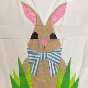 Op de afbeelding: Een stoffen konijn met een lichtbruin gezicht, zwarte ogen en een roze neus. Het konijn heeft roze en wit patroon oren en een blauw-wit gestreepte vlinderdas. Groen gras staat onderaan, de achtergrond is wit.