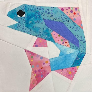 May include: A patchwork quilt block featuring a blue fish with a pink and white background. The fish is made of different fabric pieces with a swirl pattern and a black eye.