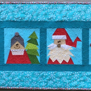 Pode incluir: Um corredor de mesa acolchoado azul com um padrão de floco de neve apresenta quatro aplicações emolduradas de personagens natalinos: um duende, uma mulher, uma árvore de Natal, Papai Noel e uma rena.