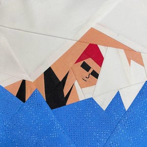 Puede incluir: Una obra de arte textil que representa a un nadador en una piscina. El nadador lleva un gorro rojo y gafas de sol negras. El agua está representada por tela azul con puntos blancos. El fondo es blanco.