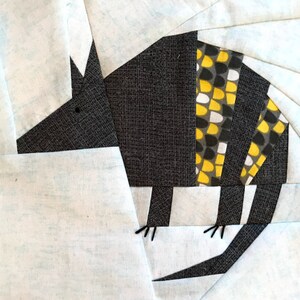 May include: A fabric art piece featuring an armadillo design. The armadillo's body is primarily dark gray with a patterned yellow and white section. The background is light blue and white, with the armadillo's legs and head visible.