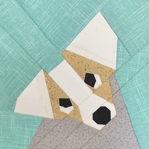 May include: A fabric patchwork dog head with a white face, tan ears, and black eyes and nose. The dog is set against a light teal background with a diagonal pattern. The patchwork design is a geometric representation of a dog.