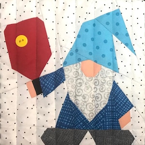 May include: A quilted fabric square featuring a gnome with a blue polka-dot hat, a white and gray swirl-patterned beard, and a blue plaid shirt. The gnome holds a red fabric shape accented with a yellow button against a white polka-dot background.