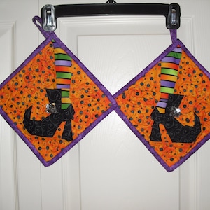 Puede incluir: Dos agarraderas de olla acolchadas de color naranja y morado con una bota de bruja negra y un diseño de pierna a rayas. Las botas tienen una gema brillante en la parte superior.