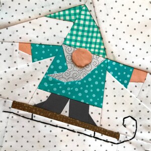May include: A patchwork quilt block featuring a gnome wearing a green and white gingham hat and a teal blue and white polka dot jacket. The gnome is standing on a brown sled with a black rope.