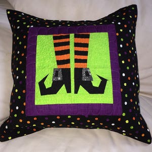 Puede incluir: Una almohada negra y verde con un borde morado y un cuadrado verde en el centro. El cuadrado presenta un par de botas de bruja negras con piernas rayadas de naranja y negro.