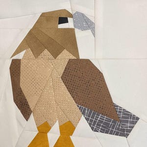 May include: A brown and tan patchwork fabric hawk with a gray beak and yellow feet. The hawk is made of different fabric patterns and is a great example of a patchwork quilt block.