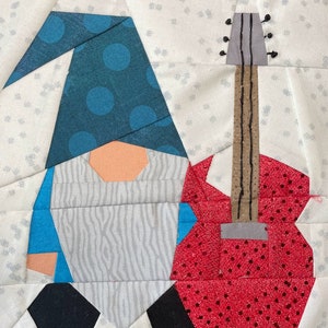 Peut inclure: Une œuvre d'art textile représentant un gnome tenant une guitare rouge. Le gnome porte un chapeau bleu à pois bleus, un nez pêche et une barbe grise. La guitare est rouge à pois noirs et un manche marron. Le fond est blanc avec un motif subtil.