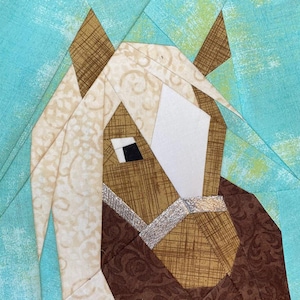 Peut inclure: Un bloc de courtepointe géométrique représentant une tête de cheval. Le cheval a un visage à motifs marron et beige, un museau blanc et un œil noir. La crinière est de couleur beige clair. Le fond est bleu clair.