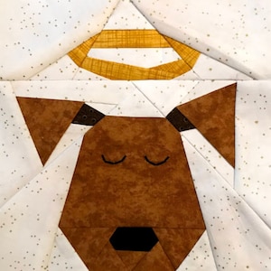 Puede incluir: Un bloque de colcha de retazos marrón y blanco con un perro durmiendo con un halo. Los ojos del perro están cerrados y tiene una nariz negra. El halo es amarillo y tiene un patrón texturizado.