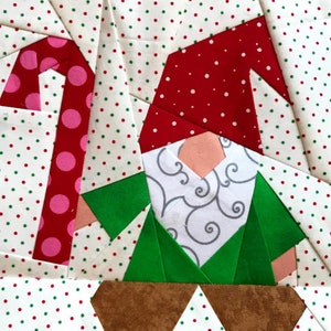 Pode incluir: Um bloco de colcha de patchwork com um gnomo com um chapéu vermelho, barba branca e jaqueta verde. O gnomo está segurando um bastão de doces rosa e branco. O fundo é um tecido branco com bolinhas vermelhas e verdes.