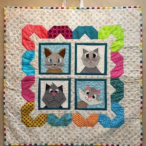 Może przedstawiać: Biały pikowany wisiorek ścienny z kolorową ramką i czterema oprawionymi obrazami kotów. Koty są w różnych kolorach i mają różne wyrażenia twarzy. Ramka wykonana jest z różnych geometrycznych kształtów i kolorów, w tym czerwonego, różowego, niebieskiego, zielonego i żółtego.