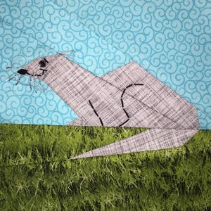 May include: A gray fabric appliqué of an otter lying on a green fabric background with a blue fabric background with a swirl pattern behind the otter.