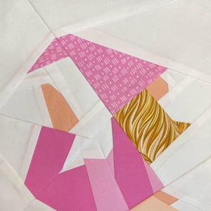May include: A patchwork quilt block featuring a pink, white, and gold fabric design. The block is made up of various geometric shapes and patterns.
